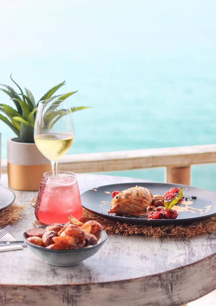 Une assiette remplie de fruits de mer, un cocktail rouge et un verre de vin blanc sur une table avec vue sur la mer des Caraïbes