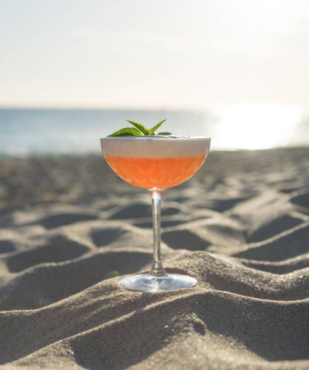 Un cocktail fruitée déposé sur le sable d'une plage avec la mer en arrière-plan.