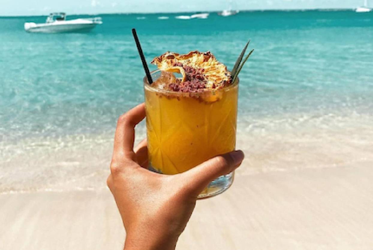 Une main tient un cocktail fruité et orangé devant une plage de sable blanc et de l'eau turquoise