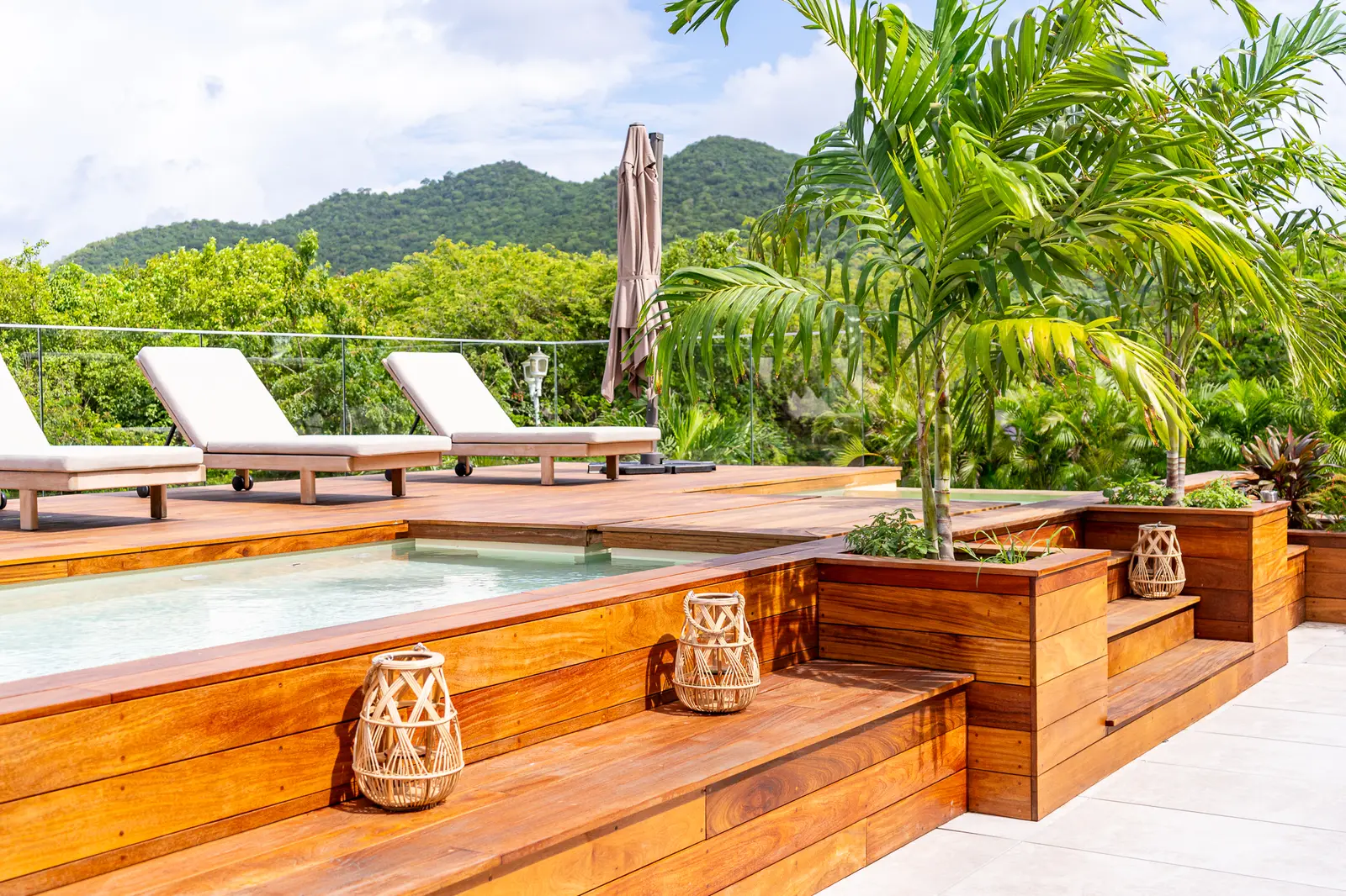 Wooden pool deck with loungers and lanterns, green hills behind.